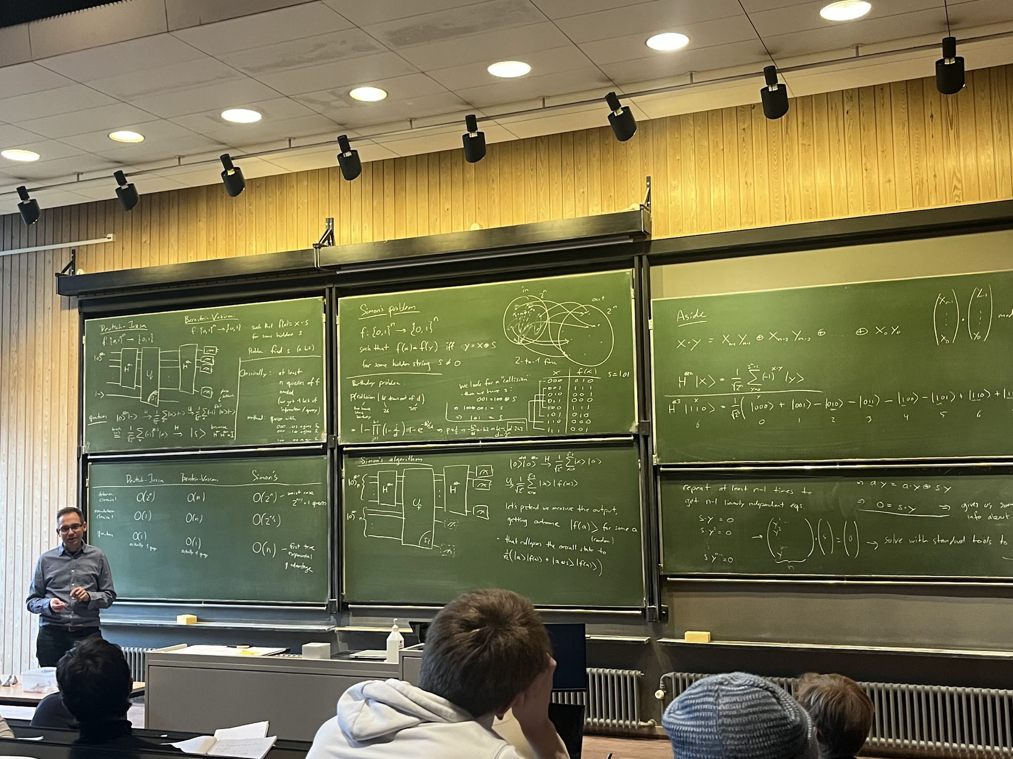 A DTU lecture hall with quantum-algorithm derivations (Deutsch–Jozsa, Simon's) across green chalkboards