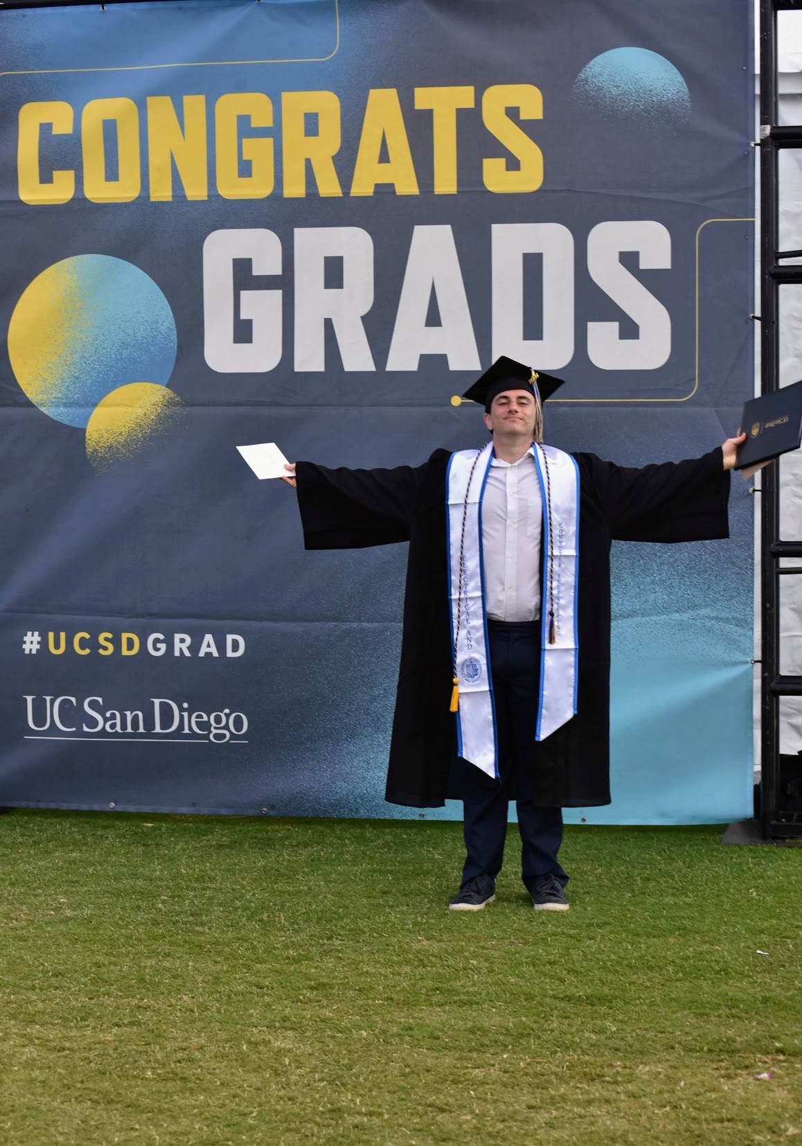 David in cap and gown before a UC San Diego “Congrats Grads” banner